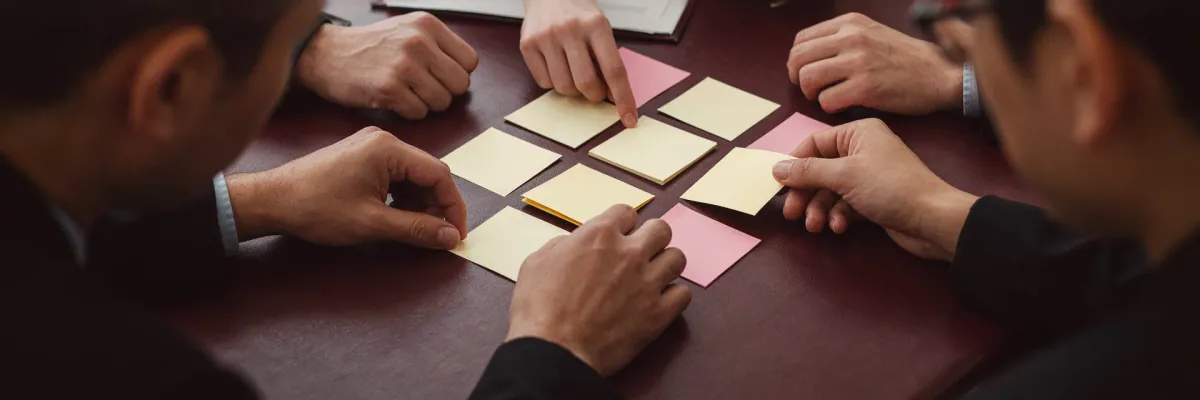Two colleagues arranging blank coloured sticky notes into a workflow grid on a desk.